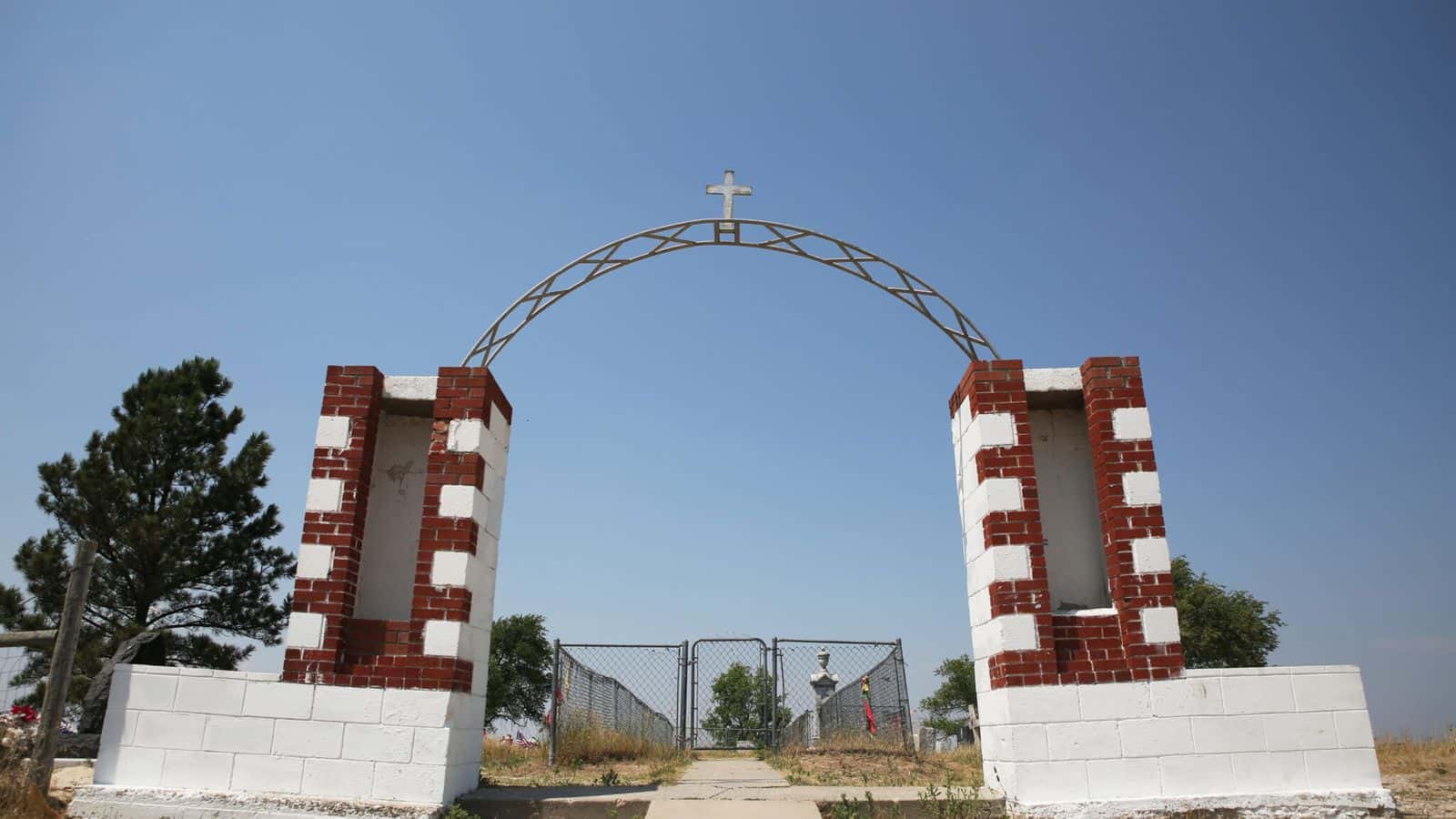 An arch topped with a cross marks the entrance to the Wounded Knee Memorial and cemetery on the Pine Ridge Reservation on June 30, 2024. (Makenzie Huber/South Dakota Searchlight)
