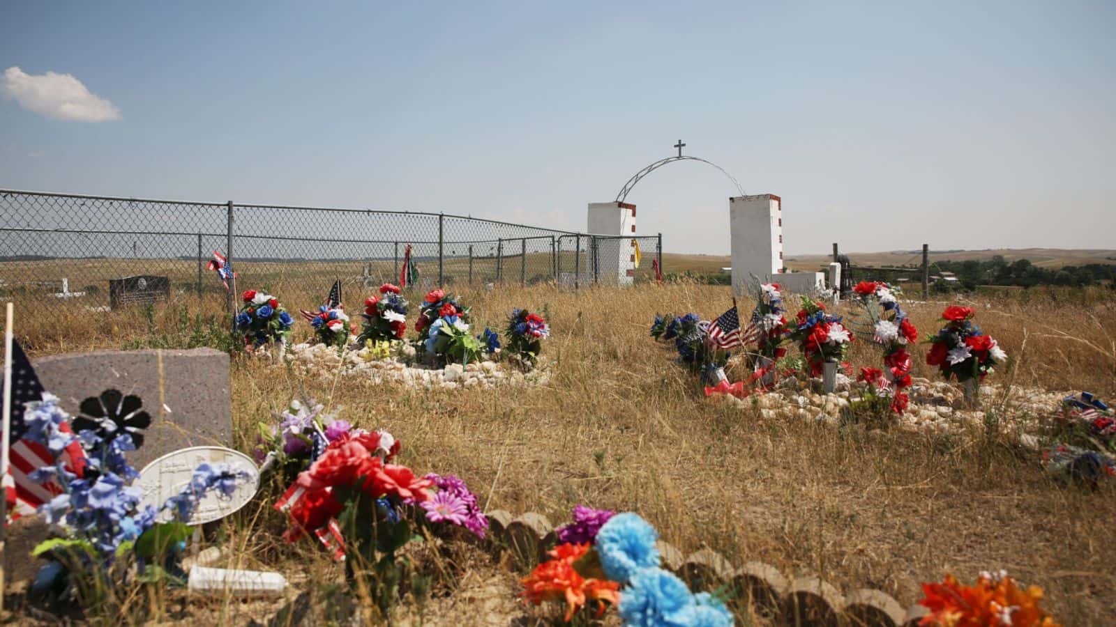 Plastic flowers and American flags are placed atop graves at the Wounded Knee Memorial and cemetery on June 30, 2024. (Makenzie Huber/South Dakota Searchlight)