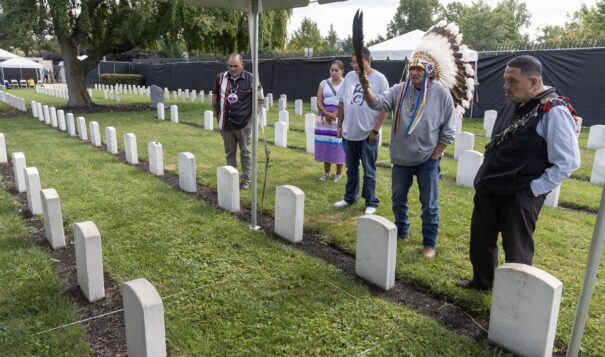 A group of Oglala Sioux gather by the grave of James Cornman (at lower left) at the Carlisle Barracks Post Cemetery in Pennsylvania. Three Oglala students who died at Carlisle - Fannie Charging Shield , Samuel Flying Horse, and James Cornman - were among 11 Native youths disinterred from the Carlisle cemetery starting Sept. 6, 2024, to be repatriated to their homelands. Pictured are, from left, Justin Pourier, Anna Diaz, Steve Durbay Jr., Steve Durbay Sr., and Jerome LeBeaux. (Photo by Charles Fox, special to ICT)